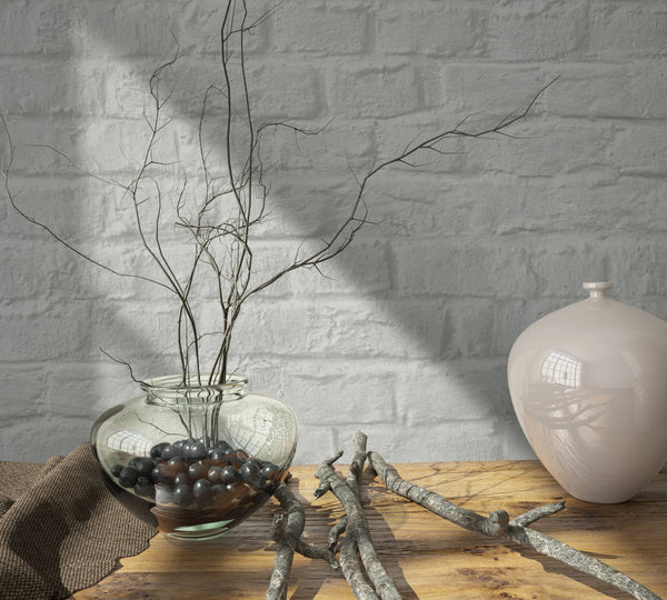 Marburg Loft Superior 34166 Light gray faux brick wallpaper behind a wooden tabletop styled with a clear glass vase holding twigs and dark decorative stones, loose branches, a brown cloth, and a glossy ceramic vase.
