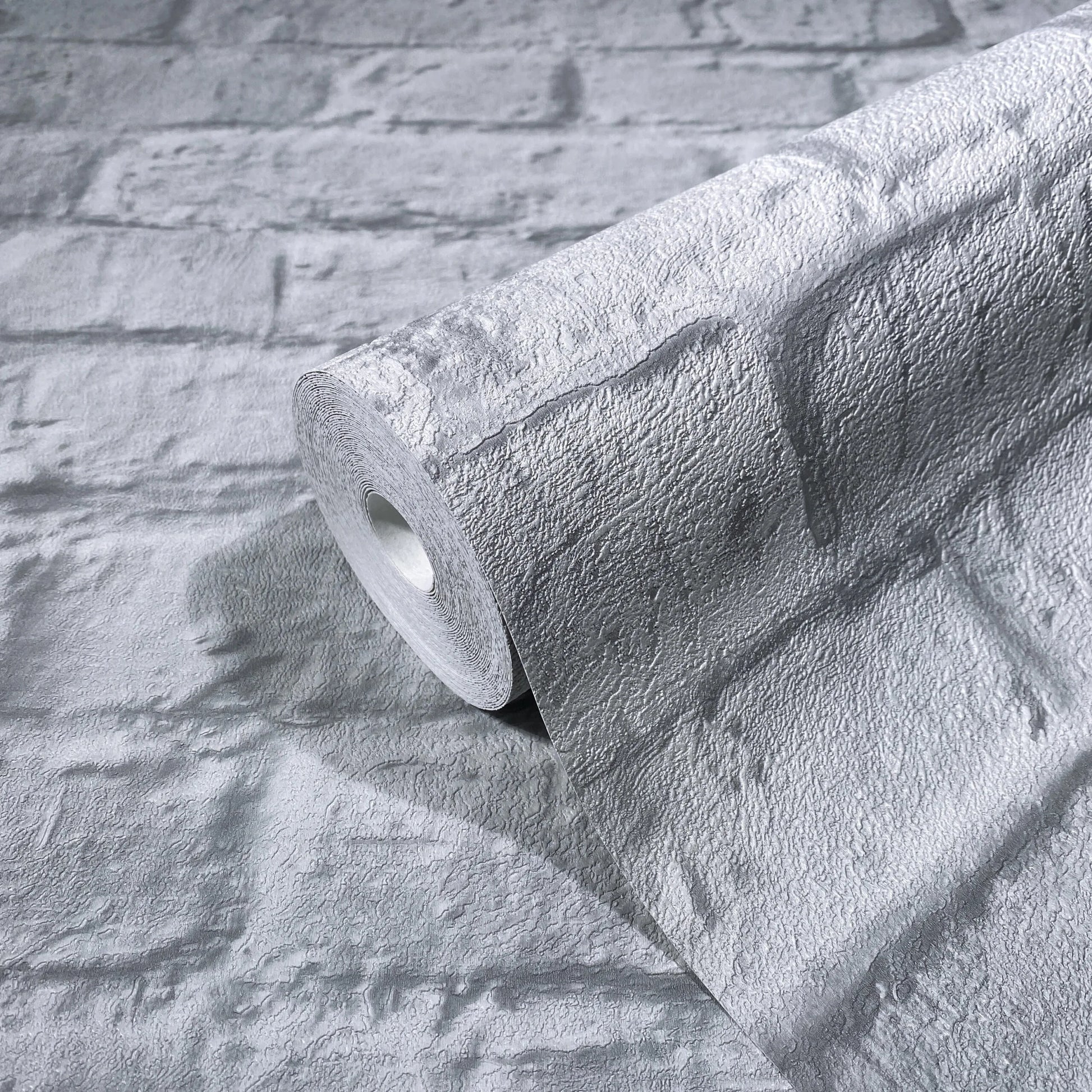 Marburg Loft Superior 34167 Close-up of a wallpaper roll with a gray textured brick pattern resting on a matching unrolled sheet.