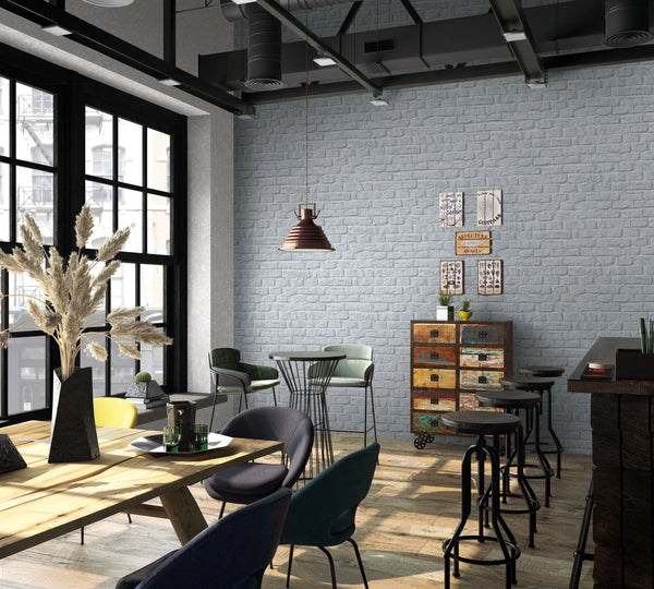 Marburg Loft Superior 34167 Gray brick-effect wallpaper covers a large wall in an industrial-style cafe with bar stools, a dining table and chairs, a pendant light, large windows, and a colorful chest of drawers.