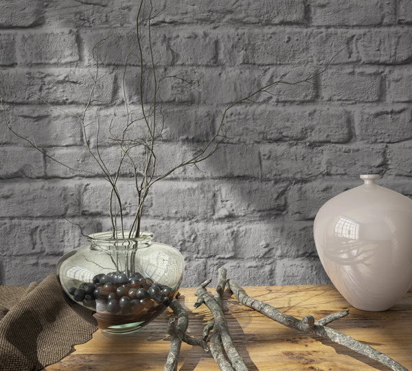 Marburg Loft Superior 34168 Styled close-up of gray brick-effect wallpaper behind a wooden tabletop with a clear glass vase holding branches and pebbles, a light ceramic vase, and scattered twigs.