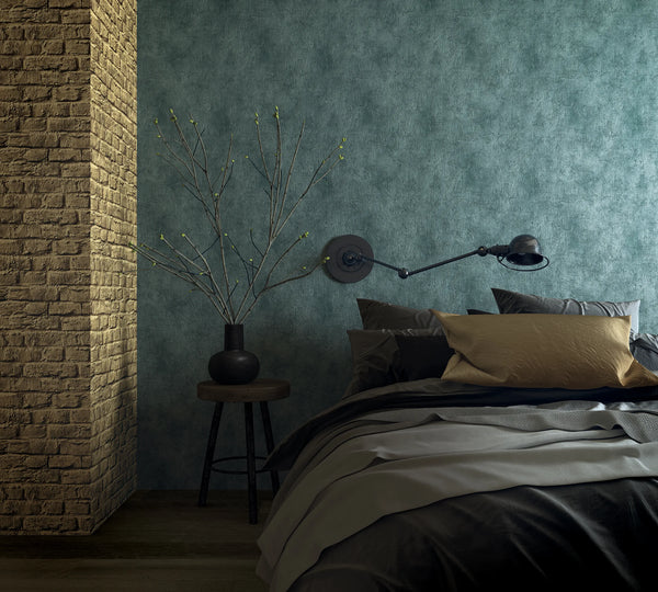 Marburg Loft Superior 34169 Blue-green mottled textured-look wallpaper behind a bed, with a swing-arm wall lamp, a stool holding a black vase with branches, and a brick column visible.