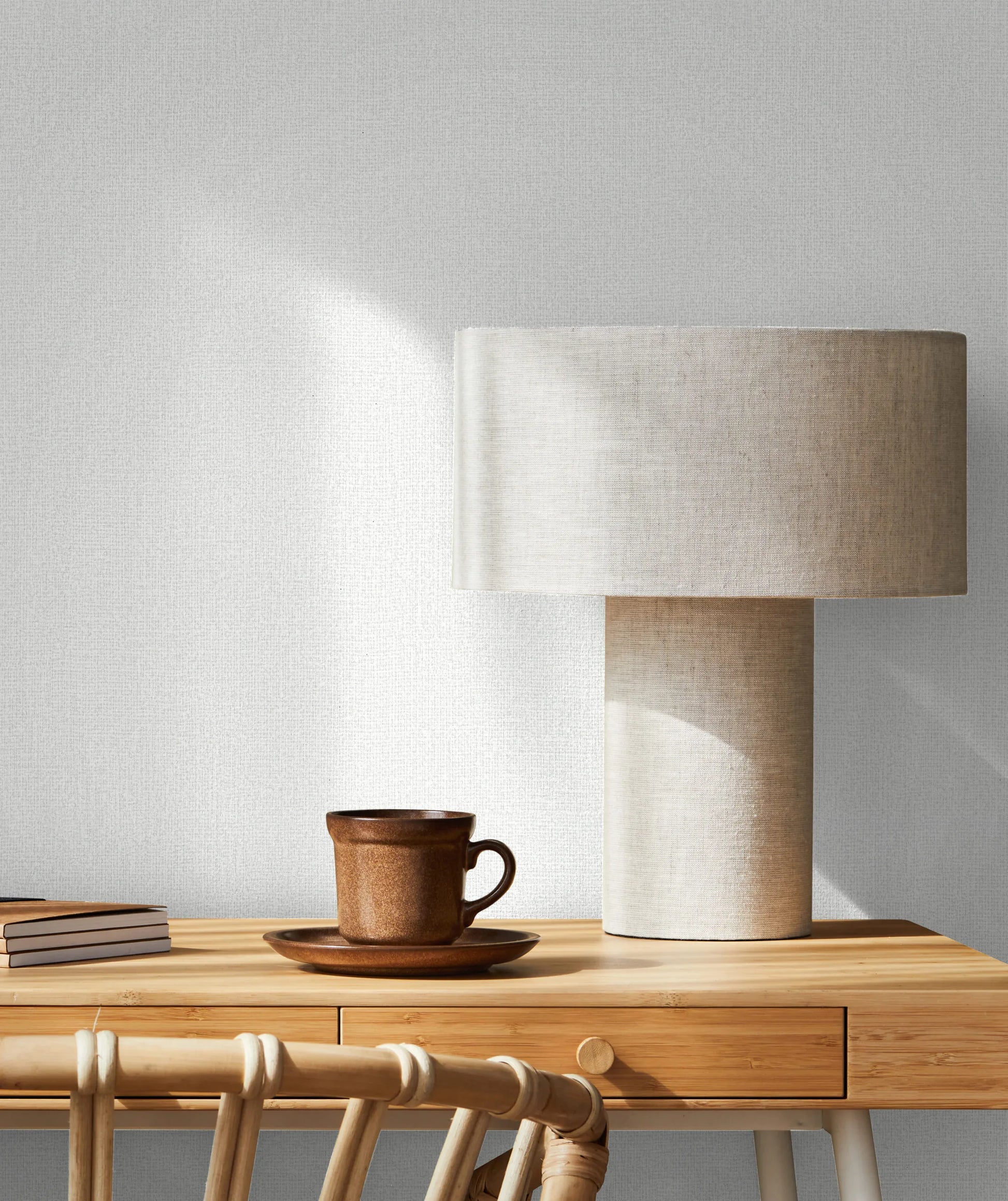 Marburg Loft Superior 34171 Light textured white wallpaper with a fine linen-like weave behind a wooden desk vignette featuring a fabric-shaded table lamp, a brown cup and saucer, stacked books, and a rattan chair.