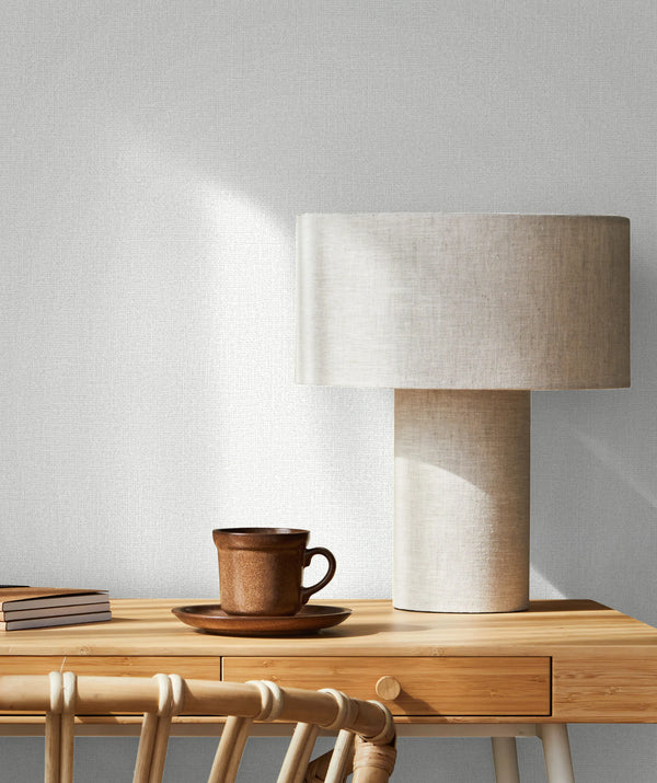 Marburg Loft Superior 34171 Light textured white wallpaper with a fine linen-like weave behind a wooden desk vignette featuring a fabric-shaded table lamp, a brown cup and saucer, stacked books, and a rattan chair.