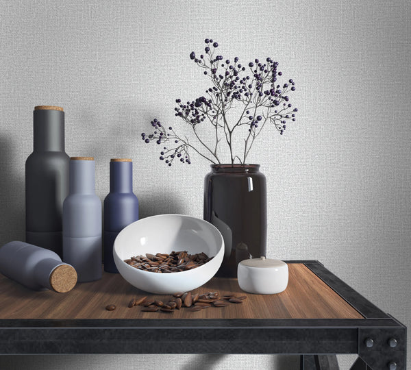 Marburg Loft Superior 34172 Light gray textured wallpaper with a fine woven look behind a tabletop vignette featuring matte bottles with cork stoppers, a white bowl holding brown seeds, a small lidded jar, and a dark glass vase with berry branches on a wood-and-metal table.