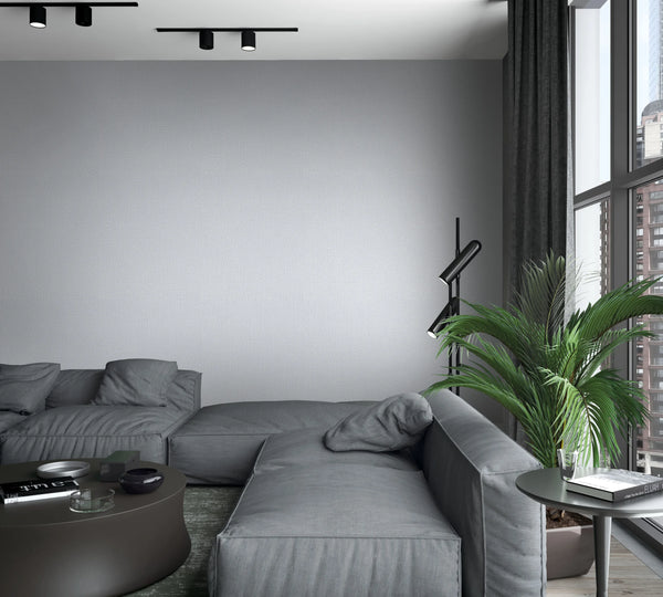 Marburg Loft Superior 34173 Light gray textured wallpaper on a feature wall behind a modular gray sofa in a modern living room, with a black floor lamp, potted palm, round coffee table, side table, curtains, and a large window.