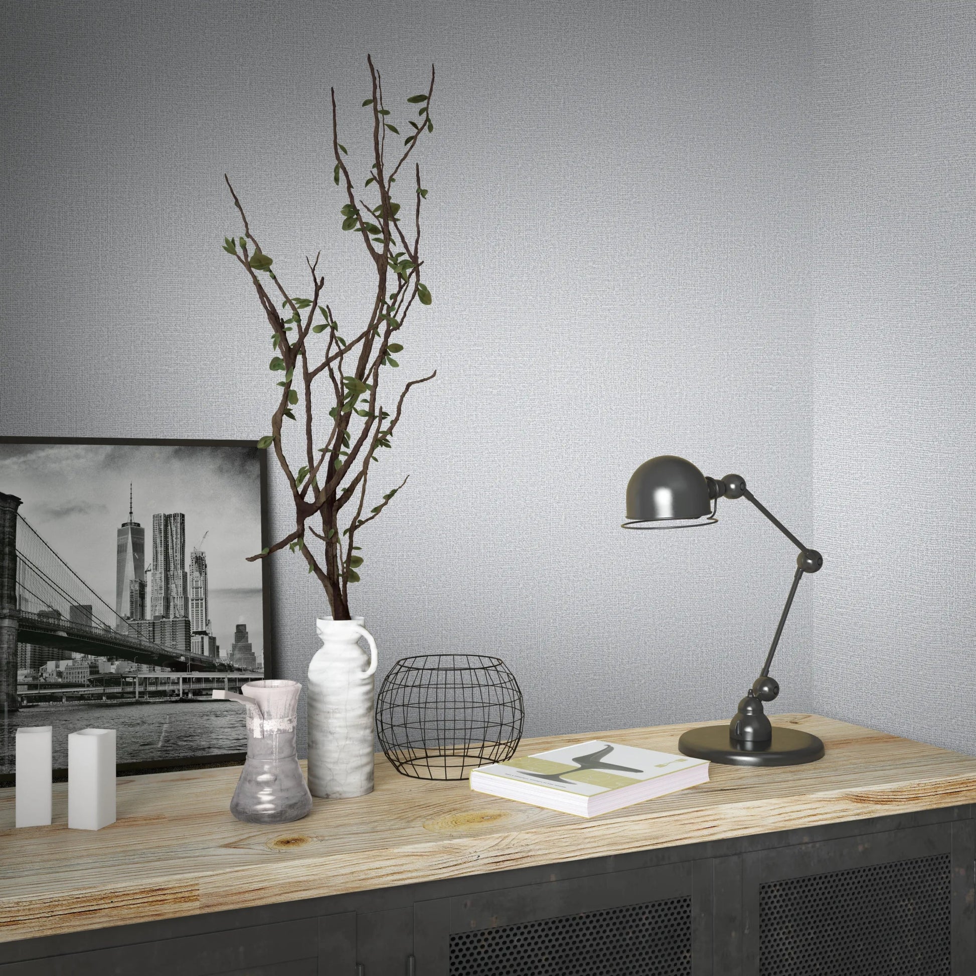 Marburg Loft Superior 34173 Light gray textured wallpaper covers two walls meeting in a corner behind a wood-top cabinet styled with a black task lamp, a framed city photograph, a white vase with branches, a wire basket, a small vase, white decorative blocks, and books.