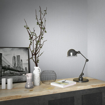 Marburg Loft Superior 34173 Light gray textured wallpaper covers two walls meeting in a corner behind a wood-top cabinet styled with a black task lamp, a framed city photograph, a white vase with branches, a wire basket, a small vase, white decorative blocks, and books.