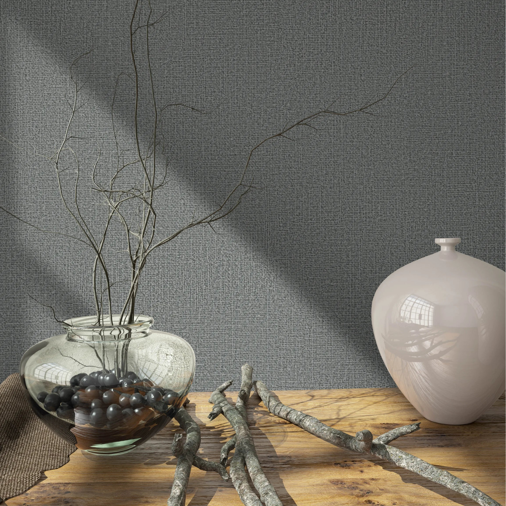 Marburg Loft Superior 34174 Close-up of gray textured wallpaper behind a wood tabletop styled with a clear glass vase holding branches, loose twigs, and a pale ceramic vase.