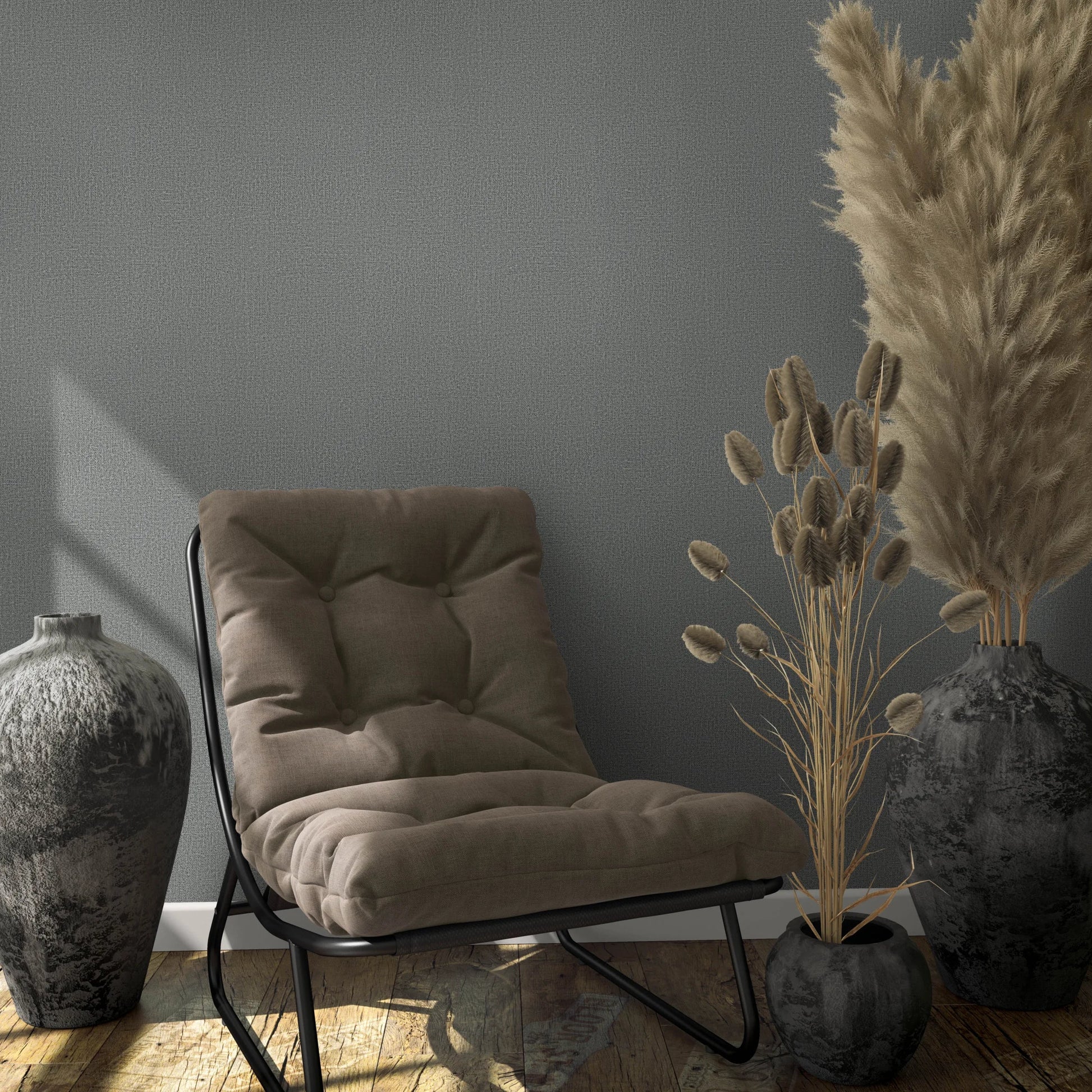 Marburg Loft Superior 34174 Gray textured wallpaper behind a cushioned lounge chair with two large dark vases and a small pot holding dried grasses on a wood floor, with windowlight shadows on the wall.
