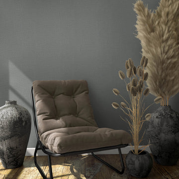 Marburg Loft Superior 34174 Gray textured wallpaper behind a cushioned lounge chair with two large dark vases and a small pot holding dried grasses on a wood floor, with windowlight shadows on the wall.