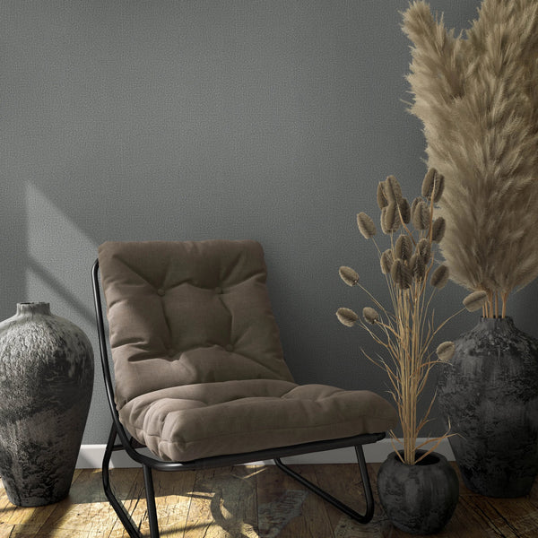 Marburg Loft Superior 34174 Gray textured wallpaper behind a cushioned lounge chair with two large dark vases and a small pot holding dried grasses on a wood floor, with windowlight shadows on the wall.