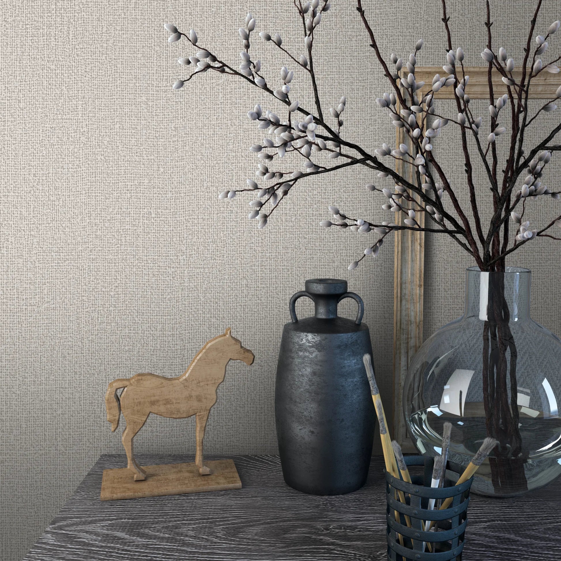 Marburg Loft Superior 34176 Textured light beige wallpaper behind a styled tabletop with a clear glass vase of branches, a dark vase, a wooden horse figurine, a wooden frame, and paintbrushes in a holder.