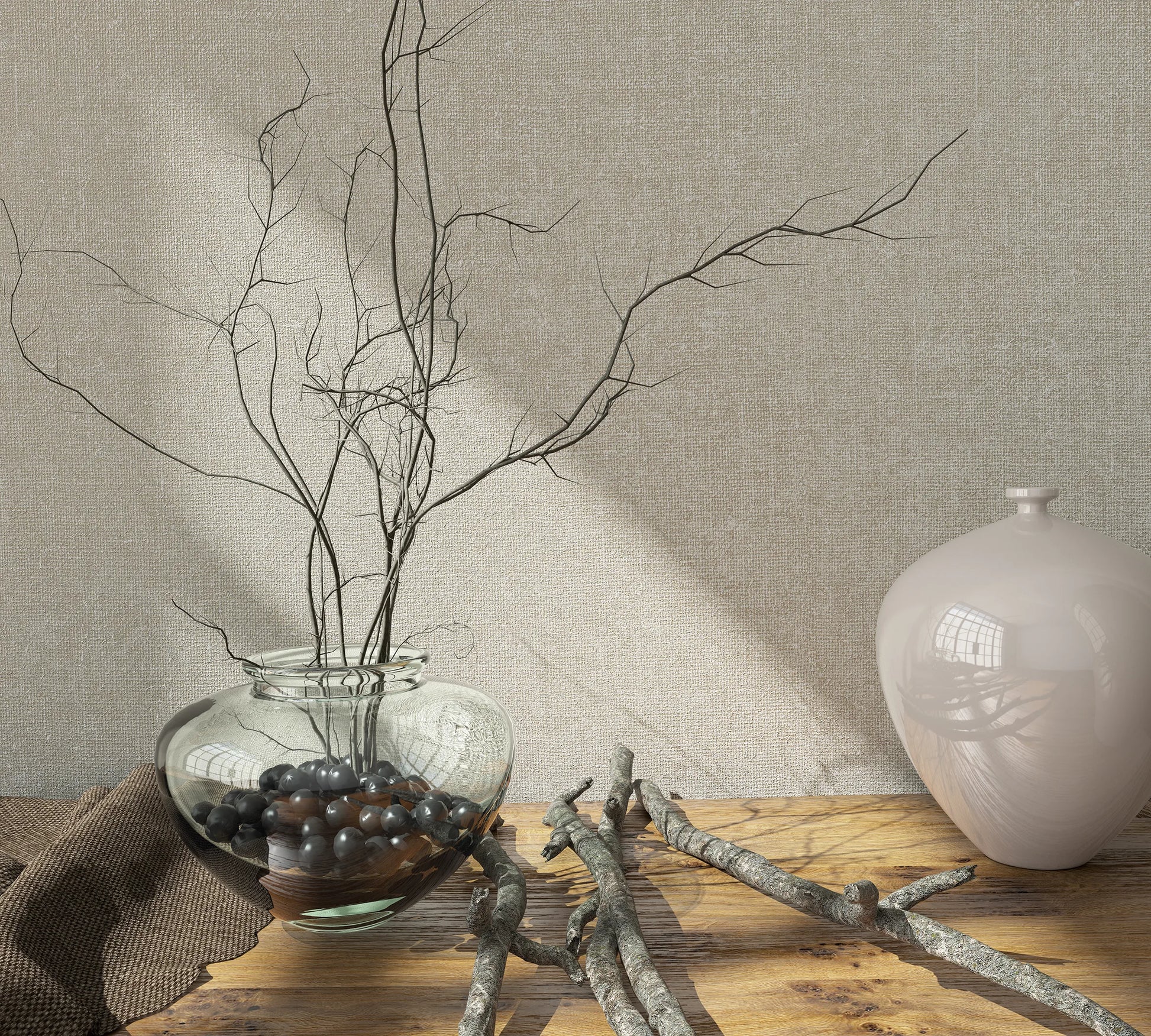 Marburg Loft Superior 34177 Beige textured wallpaper behind a wood tabletop styled with a clear glass vase holding bare branches and dark stones, a glossy pale ceramic vase, and scattered twigs in soft light.