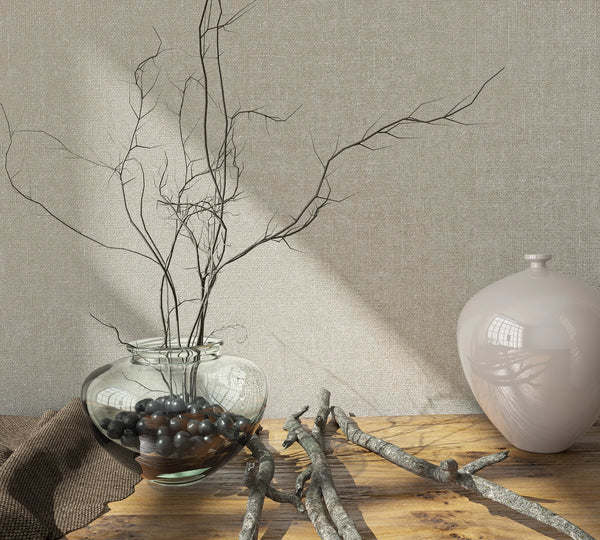 Marburg Loft Superior 34177 Beige textured wallpaper behind a wood tabletop styled with a clear glass vase holding bare branches and dark stones, a glossy pale ceramic vase, and scattered twigs in soft light.