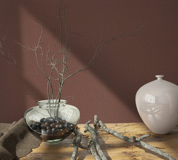Marburg Loft Superior 34179 Reddish-brown textured wallpaper behind a wooden surface styled with a clear glass bowl filled with dark stones and branches, a white ceramic vase, and scattered twigs.