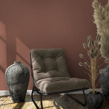Marburg Loft Superior 34179 Reddish-brown textured wallpaper behind a cushioned lounge chair with two dark ceramic vases, one filled with pampas grass, plus a small pot with dried stems on a wooden floor.