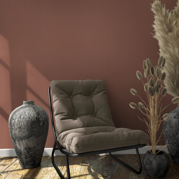 Marburg Loft Superior 34179 Reddish-brown textured wallpaper behind a cushioned lounge chair with two dark ceramic vases, one filled with pampas grass, plus a small pot with dried stems on a wooden floor.