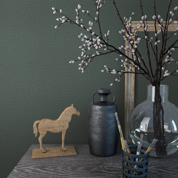 Marburg Loft Superior 34180 Close-up of green textured wallpaper behind a tabletop with a clear round glass vase of branches, a dark ceramic bottle, a small wooden horse figurine, and paintbrushes in a holder.