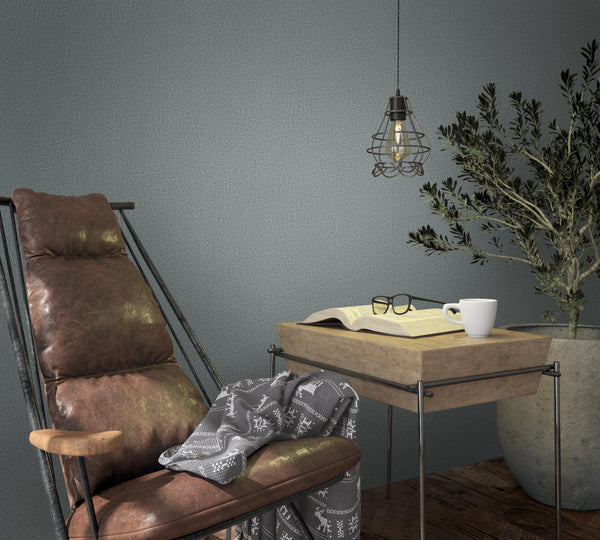 Marburg Loft Superior 34181 Muted green textured wallpaper behind a brown lounge chair, a wood side table with an open book, eyeglasses, and a white mug, a wire‑cage pendant light, and a large potted plant on a wood floor.
