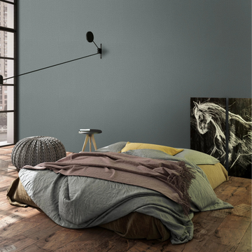 Marburg Loft Superior 34181 Textured green wallpaper on the feature wall behind a low bed in a modern bedroom, with a long-arm wall sconce, knitted pouf, small stool table, large grid window, and horse artwork panels leaning against the wall.