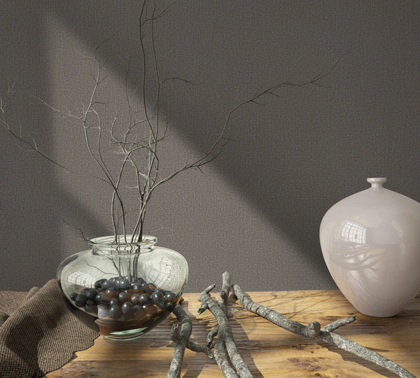 Marburg Loft Superior 34183 Textured brown wallpaper behind a wooden tabletop with a clear glass bowl filled with dark stones and twigs, loose branches, and a glossy white vase.