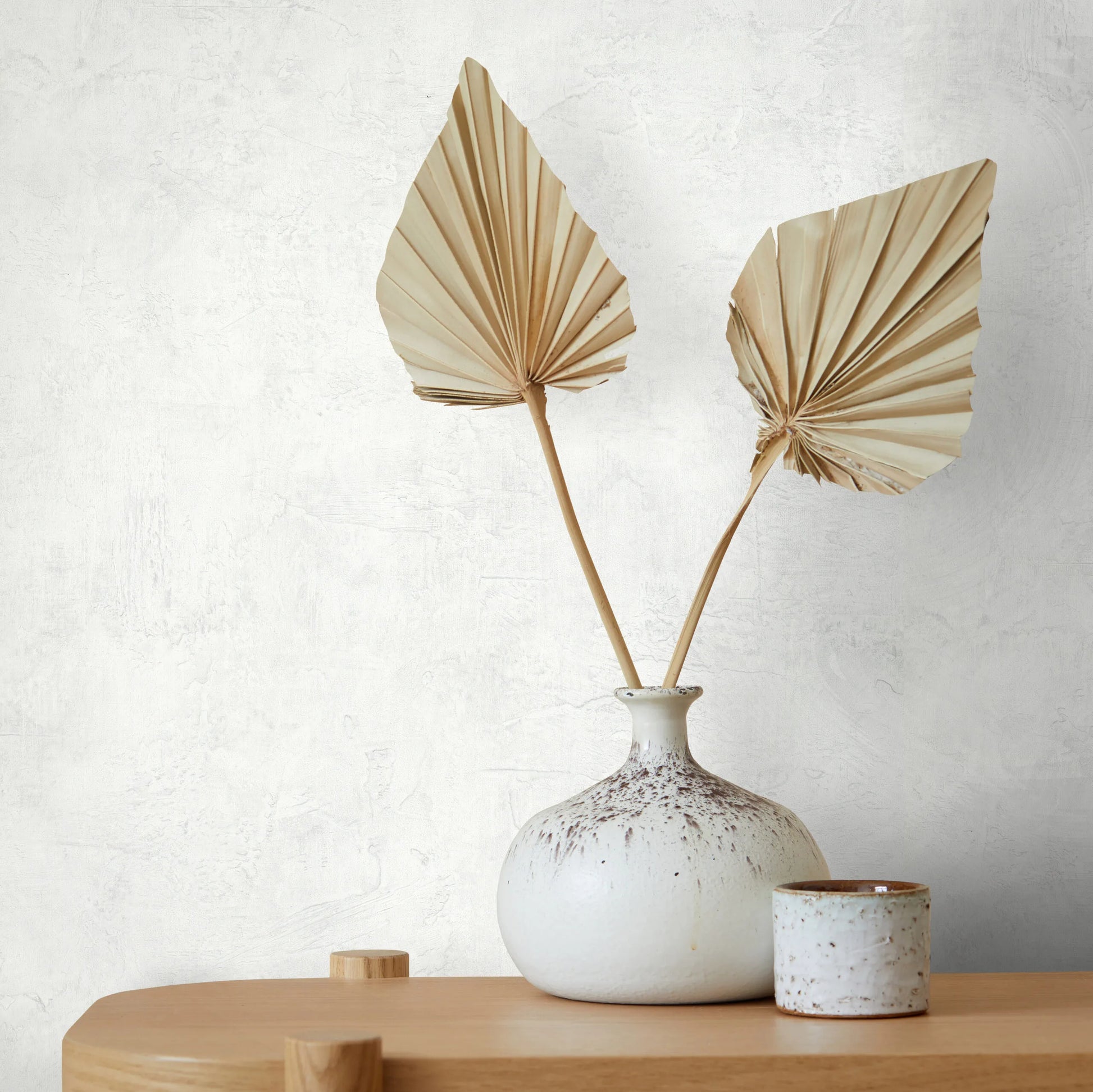 Marburg Loft Superior 34184 White plaster-effect wallpaper behind a speckled ceramic vase holding two dried palm leaves, with a small ceramic cup on a light wood surface.