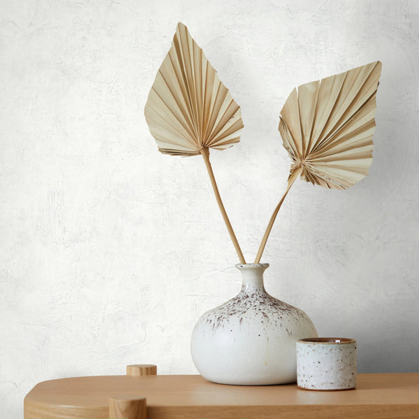 Marburg Loft Superior 34184 White plaster-effect wallpaper behind a speckled ceramic vase holding two dried palm leaves, with a small ceramic cup on a light wood surface.