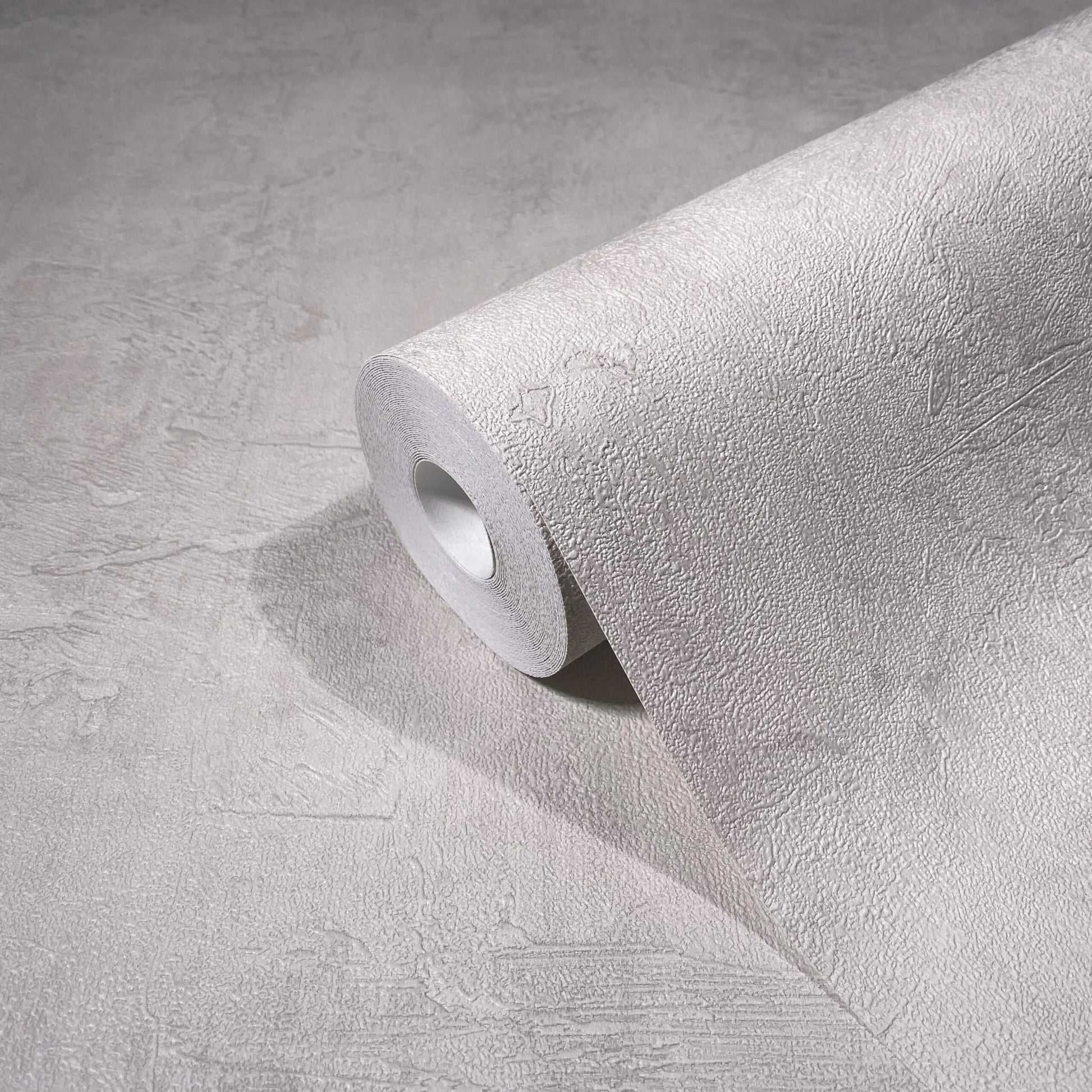 Marburg Loft Superior 34186 Angled roll of light neutral textured wallpaper with a subtle plaster effect, partially unrolled on a matching surface.