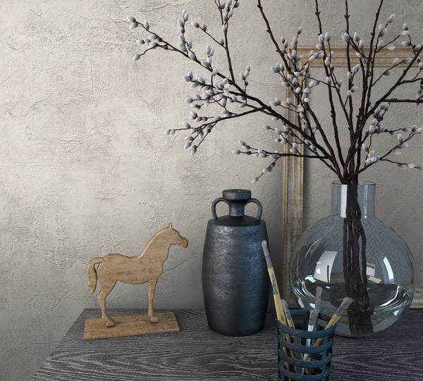 Marburg Loft Superior 34186 Close-up styled scene showing light beige textured plaster-look wallpaper behind a tabletop with a clear glass vase of pussy willow branches, a dark jug, a small wooden horse figurine, a cup with paintbrushes, and a leaning frame.