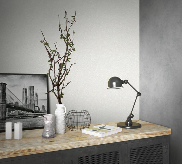 Marburg Loft Superior 34187 Light gray wallpaper with a fine, scratch-like texture is shown on a wall beside a darker gray corner. In front is a cabinet topped with a black desk lamp, a framed city photo, a white jug holding branches, a wire basket, a book, a glass vessel, and two small白.