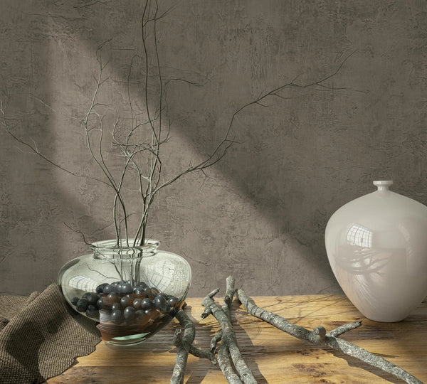 Marburg Loft Superior 34188 Textured brown wallpaper with a plaster-like look behind a wooden table styled with a clear glass vase of twigs, a white ceramic vase, loose branches, and a cloth.