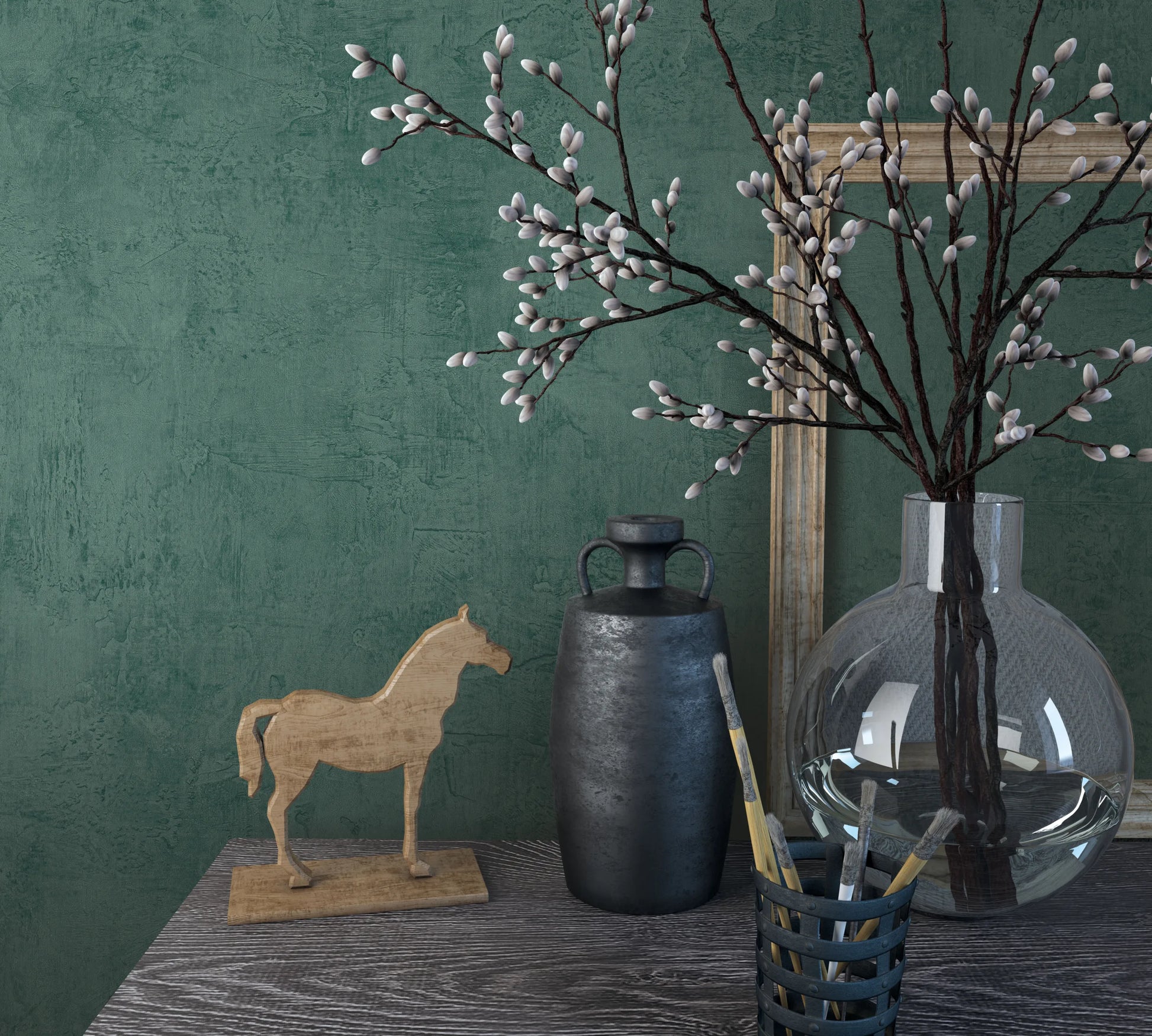 Marburg Loft Superior 34189 Green textured wallpaper with a plaster effect behind a tabletop vignette featuring a clear round glass vase of pussy willow branches, a dark jug, a wooden horse figurine, paintbrushes in a holder, and a leaning frame.