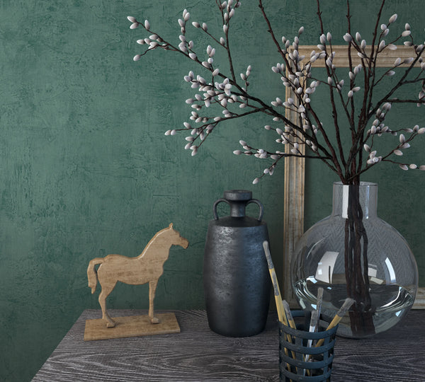 Marburg Loft Superior 34189 Green textured wallpaper with a plaster effect behind a tabletop vignette featuring a clear round glass vase of pussy willow branches, a dark jug, a wooden horse figurine, paintbrushes in a holder, and a leaning frame.