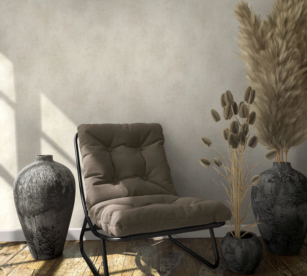 Marburg Loft Superior 34191 Subtle beige plaster‑effect wallpaper shown in a styled corner with a cushioned lounge chair, ceramic vases, dried grasses, wood floor, and window light shadows.