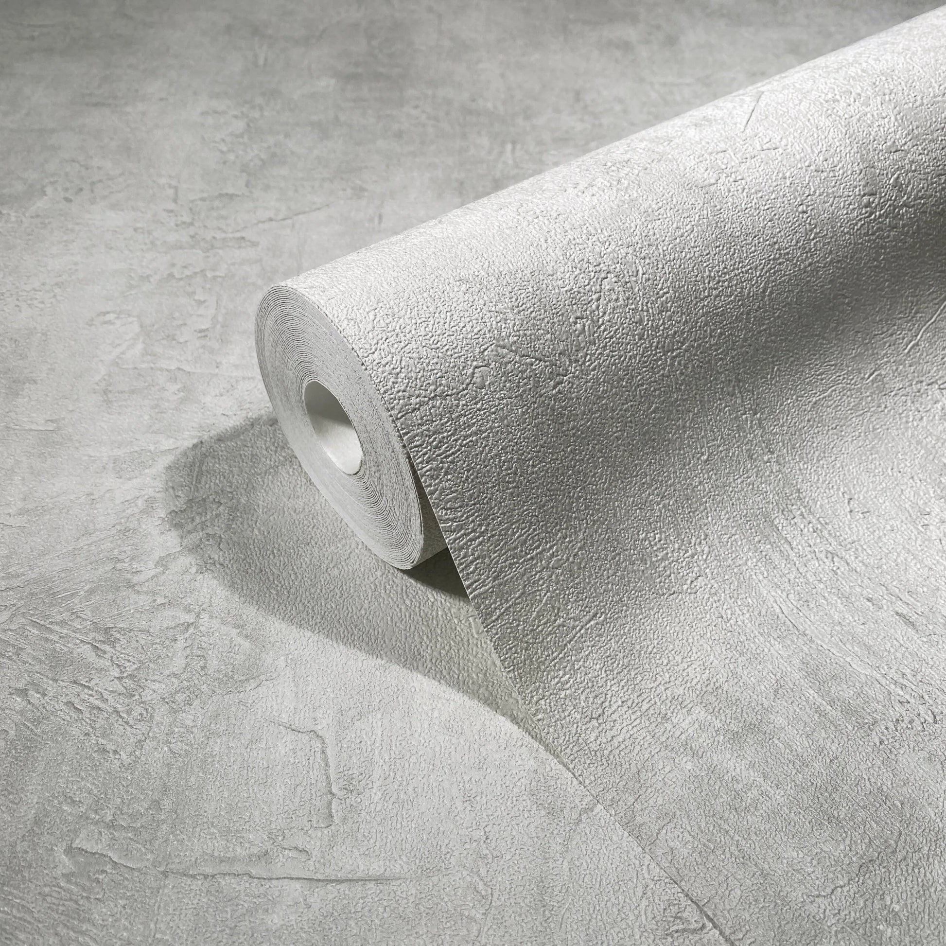 Marburg Loft Superior 34192 Close-up of a light gray textured wallpaper roll unrolling across a matching surface, showing the plaster-like pattern.