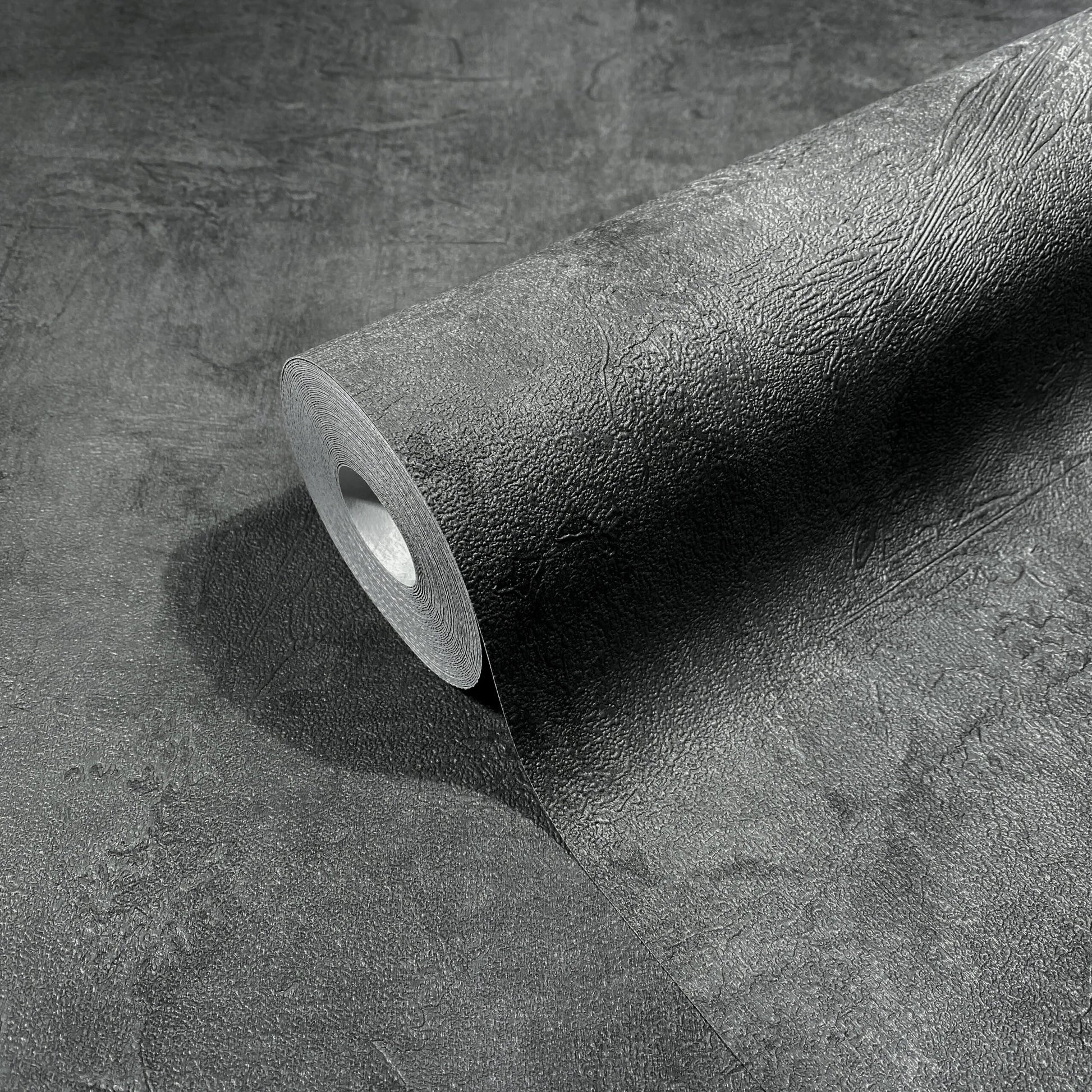Marburg Loft Superior 34193 A dark anthracite wallpaper roll partially unrolled, displaying a textured, plaster-like surface.
