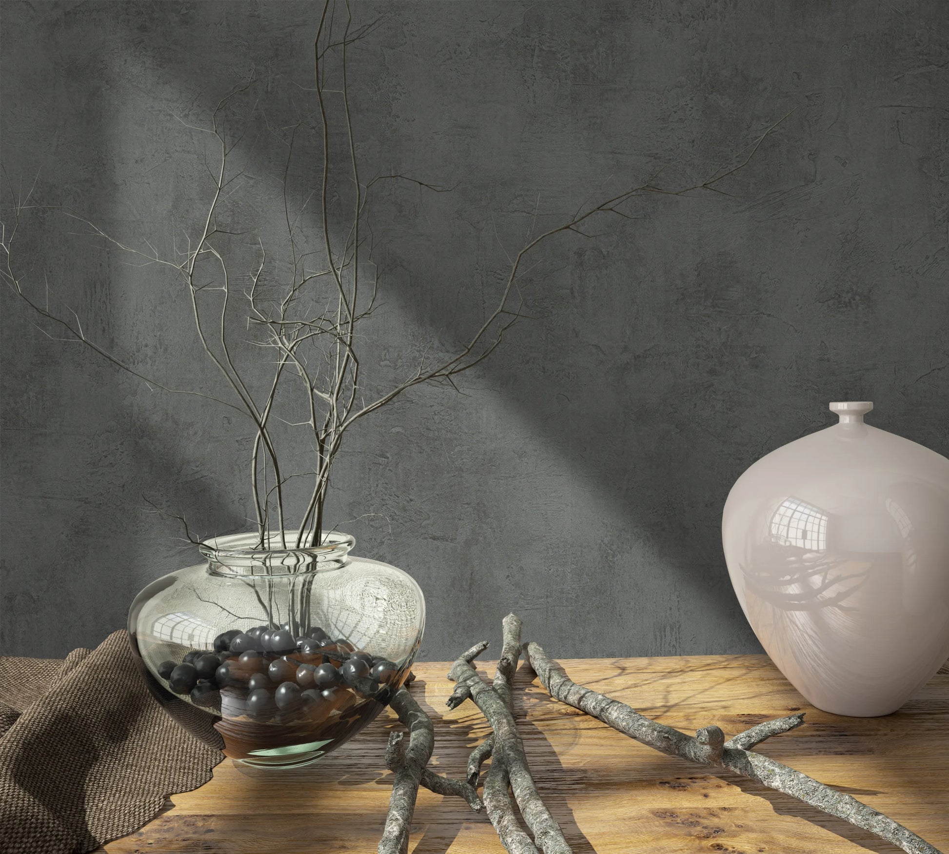 Marburg Loft Superior 34193 Dark grey concrete-look wallpaper with a mottled texture behind a wood tabletop styled with a clear glass vase of branches, dark pebbles, a light ceramic jar, loose twigs, and a brown cloth.