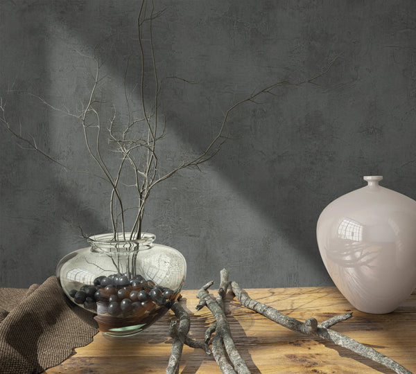 Marburg Loft Superior 34193 Dark grey concrete-look wallpaper with a mottled texture behind a wood tabletop styled with a clear glass vase of branches, dark pebbles, a light ceramic jar, loose twigs, and a brown cloth.
