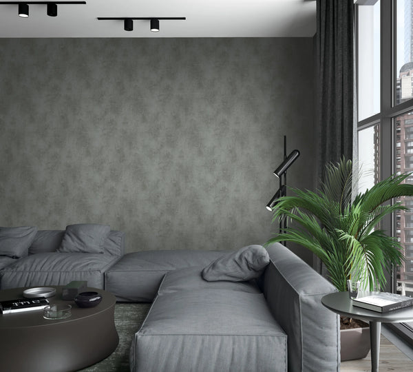Marburg Loft Superior 34193 Anthracite grey wallpaper with a soft mottled, concrete-like texture on a feature wall in a modern living room with a low grey sectional, a black floor lamp, a round coffee table, an indoor palm by tall windows, dark curtains, and ceiling track lights.