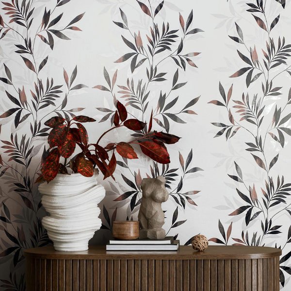 Marburg Mona 82458 Botanical wallpaper with layered leafy branches in black, gray, and muted rose on a light background, shown behind a wood console holding a sculptural white vase with red-leaf plant, a small figurine, stacked books, a small cup, and a decorative sphere.