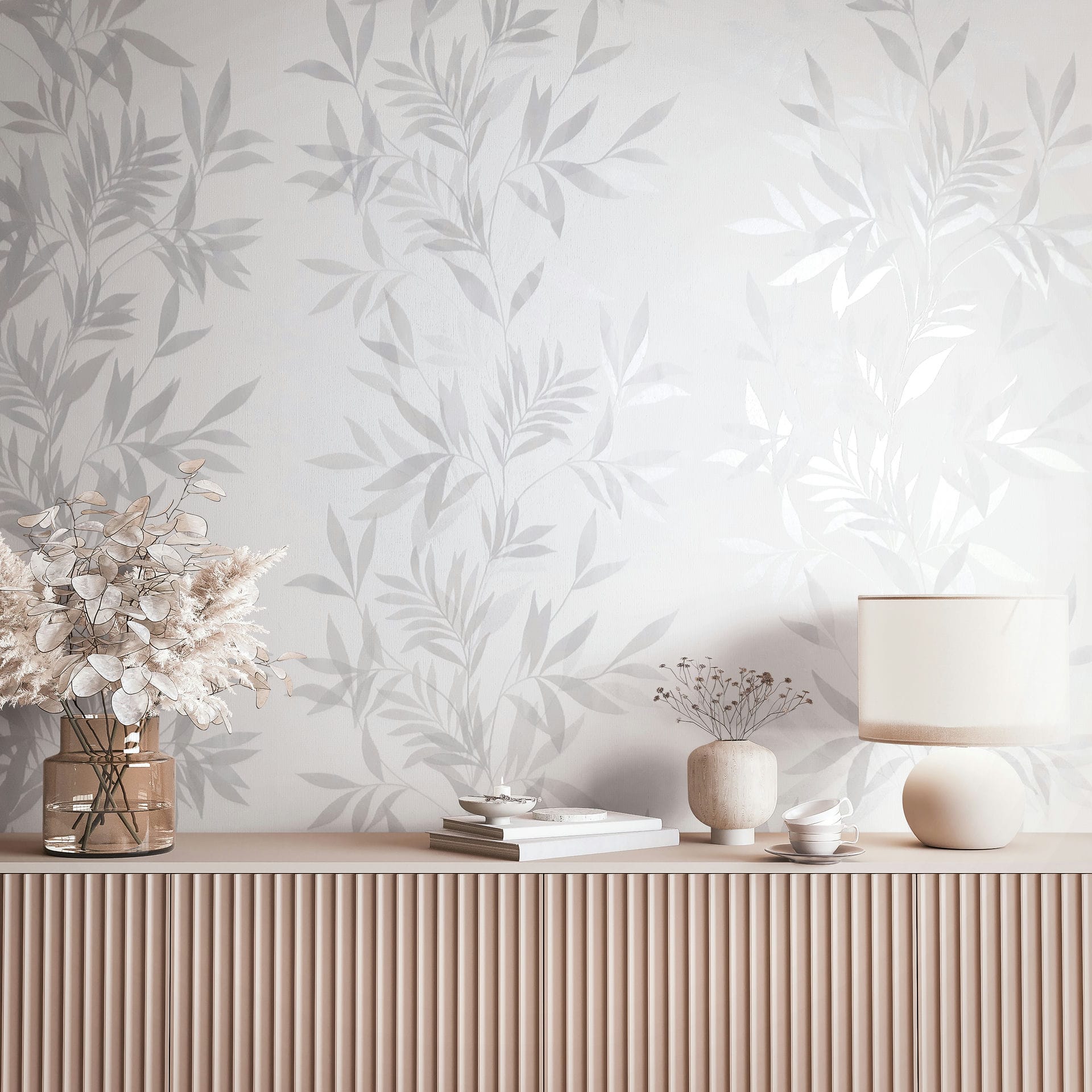 Marburg Mona 82460 Tonal gray and white botanical leaf wallpaper shown behind a fluted sideboard styled with a table lamp, a glass vase with dried foliage, a small ceramic vase with stems, stacked books, a small bowl, and a teacup with saucer.