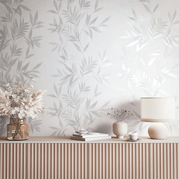 Marburg Mona 82460 Tonal gray and white botanical leaf wallpaper shown behind a fluted sideboard styled with a table lamp, a glass vase with dried foliage, a small ceramic vase with stems, stacked books, a small bowl, and a teacup with saucer.
