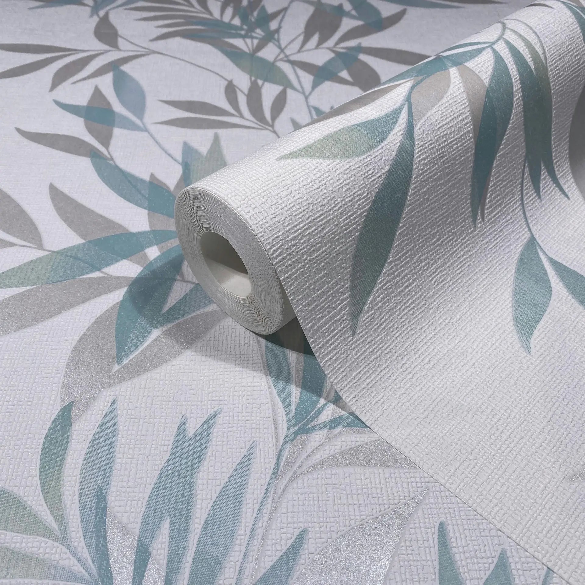 Marburg Mona 82461 Close-up of a wallpaper roll on matching unrolled wallpaper, showing a textured white surface with teal green and light gray leaf motifs.
