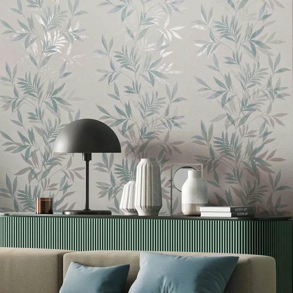 Marburg Mona 82461 Botanical leafy wallpaper in soft green on a light background behind a green ribbed console topped with a black dome table lamp, ceramic vases, and books, with part of a sofa and pillows in front.