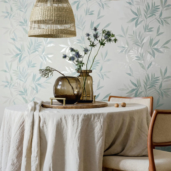 Marburg Mona 82461 Pale green and white leafy wallpaper behind a round table draped in linen, with two glass vases on a wooden tray, upholstered chairs, and a woven pendant light above.