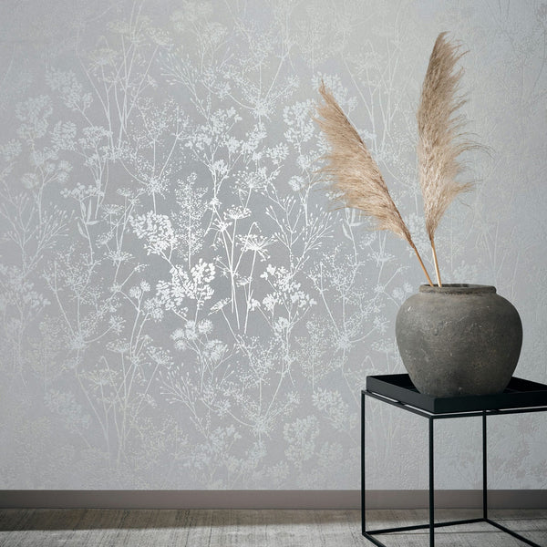 Marburg Mona 82462 Light grey wallpaper with delicate botanical silhouettes shown in a styled room scene. A large stone vase holding two pampas stems sits on a black metal side table against the wall, with baseboard and floor visible.