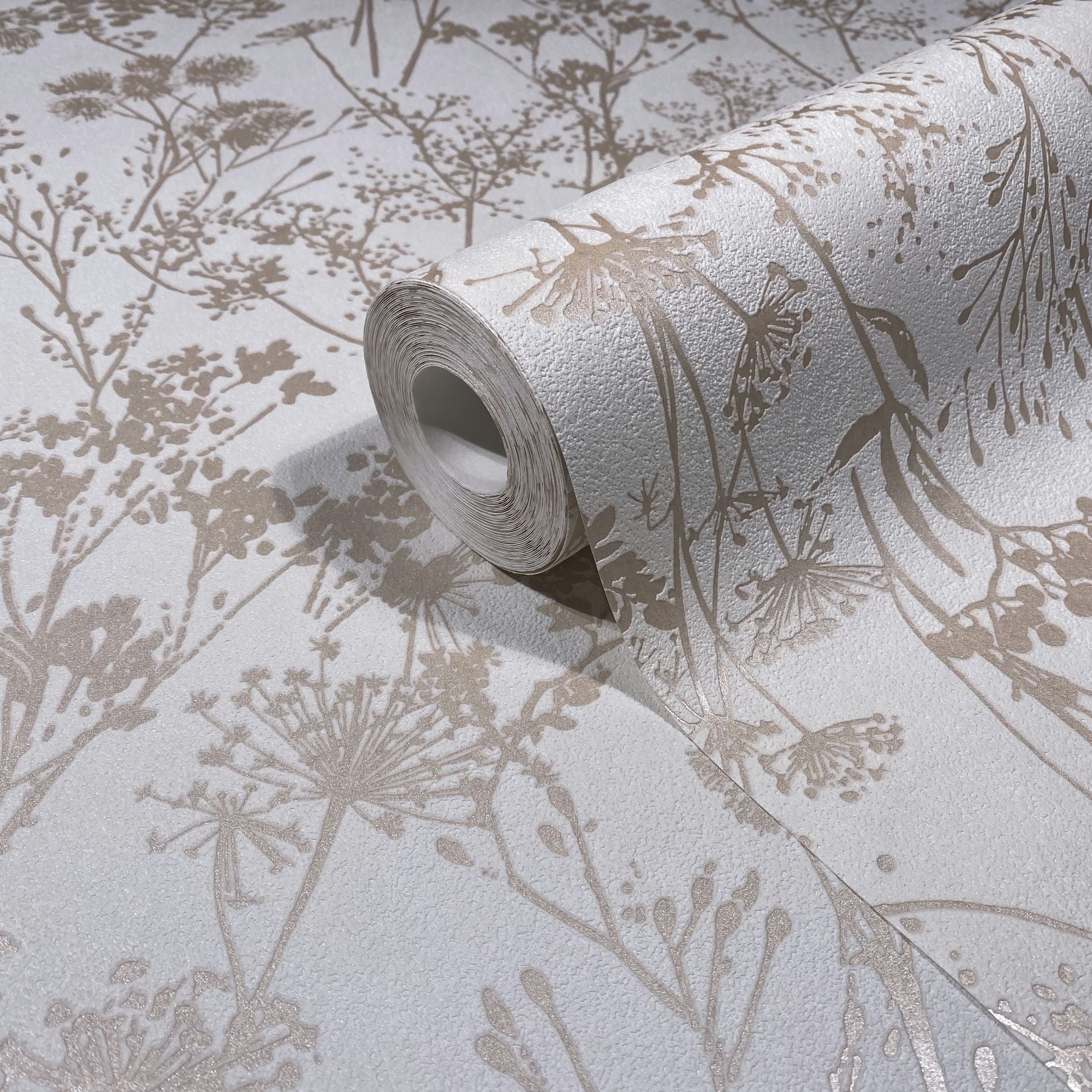 Marburg Mona 82463 Close-up of a wallpaper roll resting on matching wallpaper, showing beige botanical silhouettes of wildflowers on a textured white background.