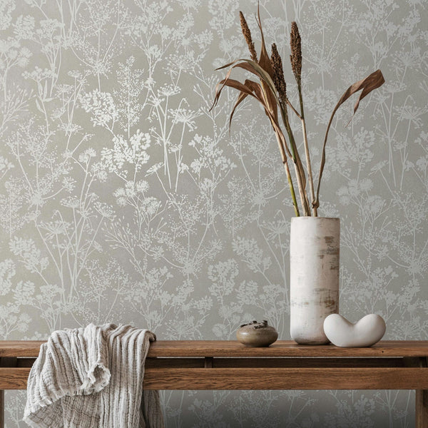 Marburg Mona 82464 Gray wallpaper with light botanical silhouettes forms the backdrop for a wooden bench holding a tall ceramic vase with dried stems, a small bowl, a sculptural white object, and a draped cloth.