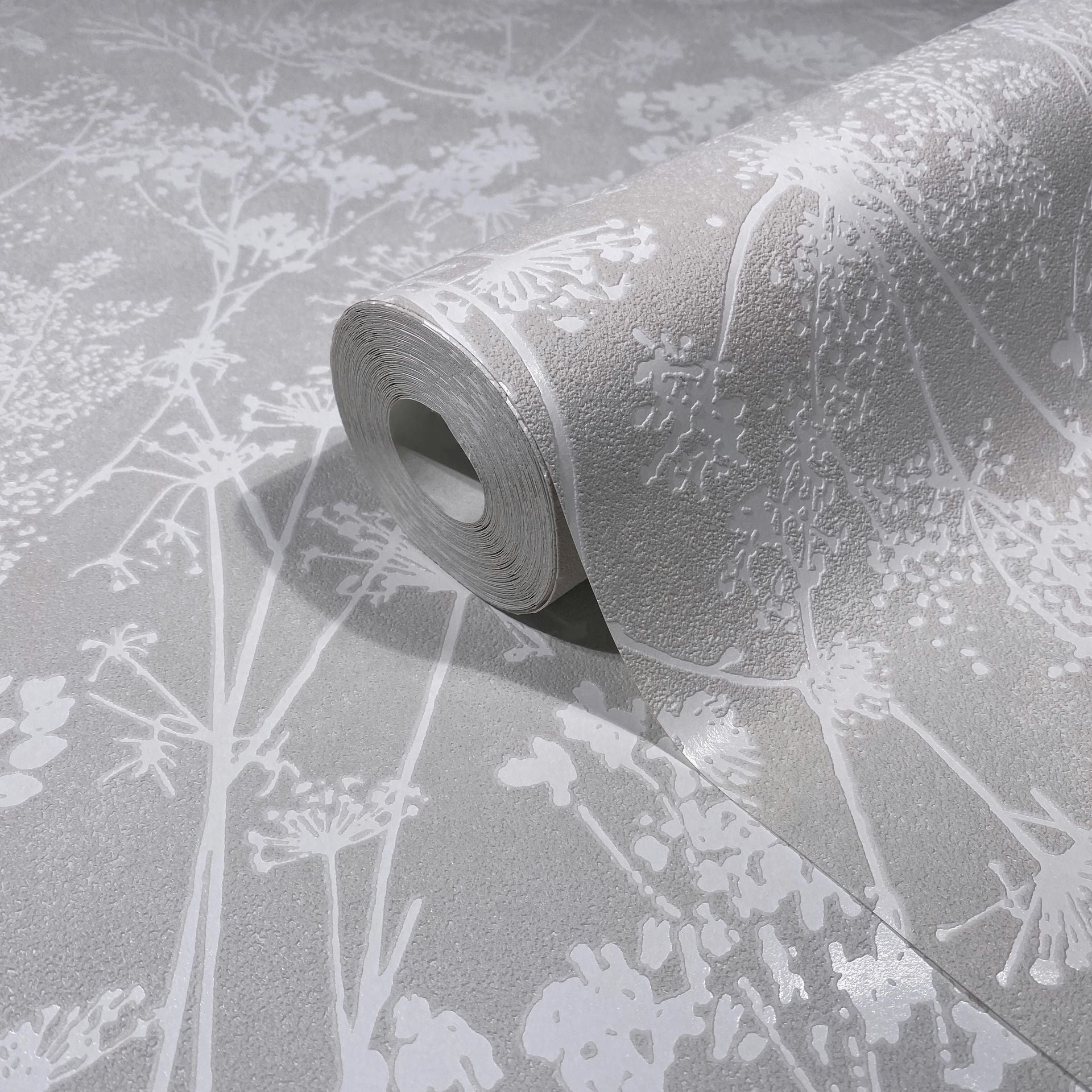 Marburg Mona 82464 A wallpaper roll partially unrolled on a matching surface, showing a light gray textured ground with glossy white botanical silhouettes.