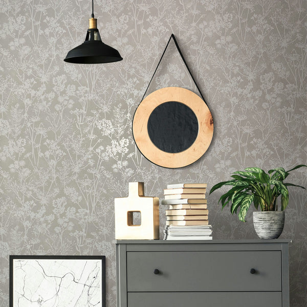 Marburg Mona 82464 Gray wallpaper with subtle botanical silhouettes is shown behind a vignette featuring a round strap-hung mirror, a black pendant light, a gray dresser with stacked books, a sculptural decor object, a framed print, and a potted plant.
